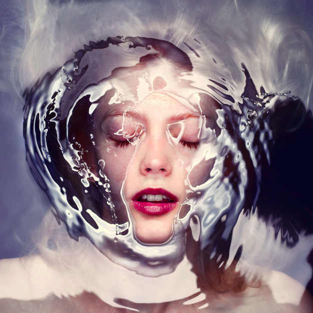 Surreal portrait of a face submerged beneath water or liquid surface, eyes closed in serene expression, surrounded by rippling distortion and refraction patterns in blue-grey and white tones; red lipstick provides a vivid accent against the…