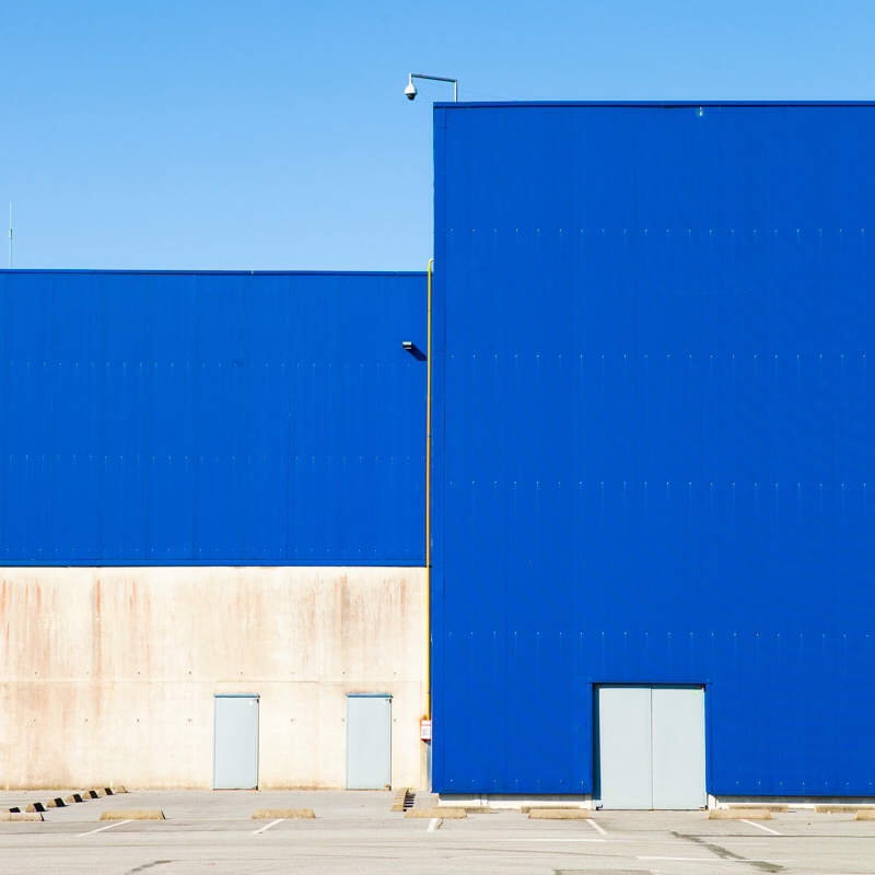Minimalist architectural photograph of an industrial building facade with bold cobalt-blue corrugated metal cladding and cream-colored concrete base, featuring geometric forms, small gray access doors, a yellow drainpipe, mounted security camera…