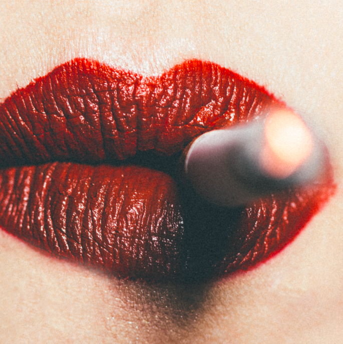 Extreme close-up macro photograph of red lipstick-painted lips with a lit cigarette between them, shot with shallow depth of field and warm intimate lighting.