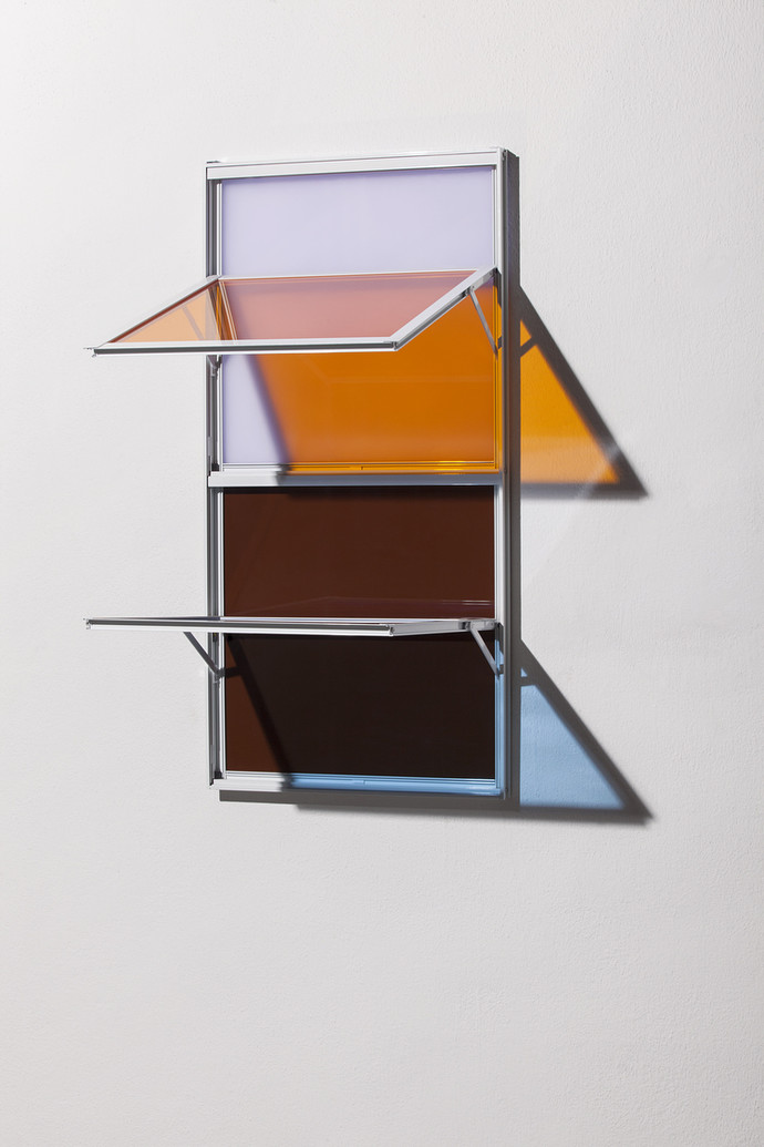A wall-mounted sculptural object made of aluminum frames and tinted glass panes in lavender, amber-orange, brown, and dark tones, with hinged glass doors that open outward.